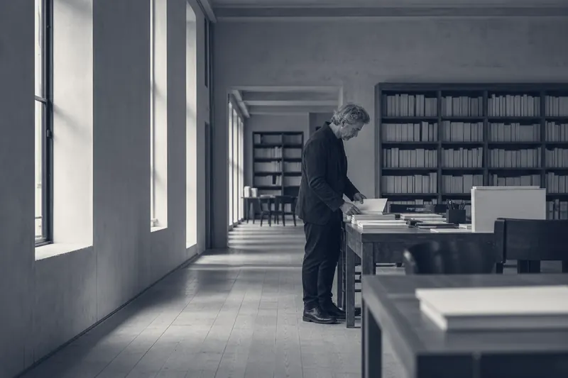 A quiet workroom with manuscripts on the desks and a man reading at the far end