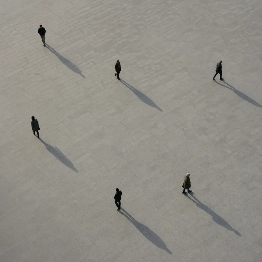 Seen from above: six people walking across a pale square, each on their own path, long shadows behind them